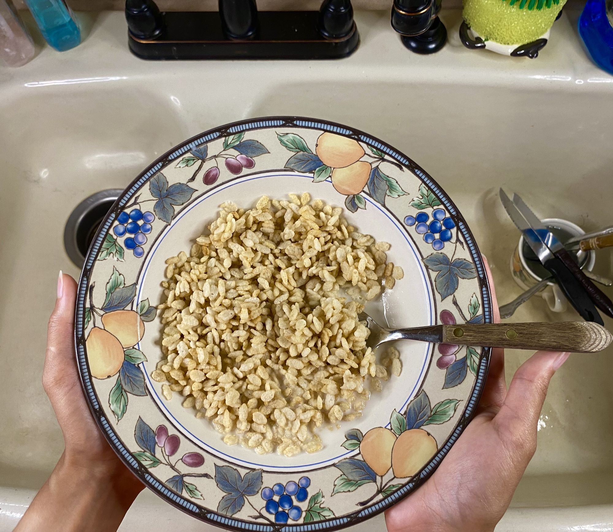A bowl of rice cereal being held over a kitchen sink with some utensils in the sink. 