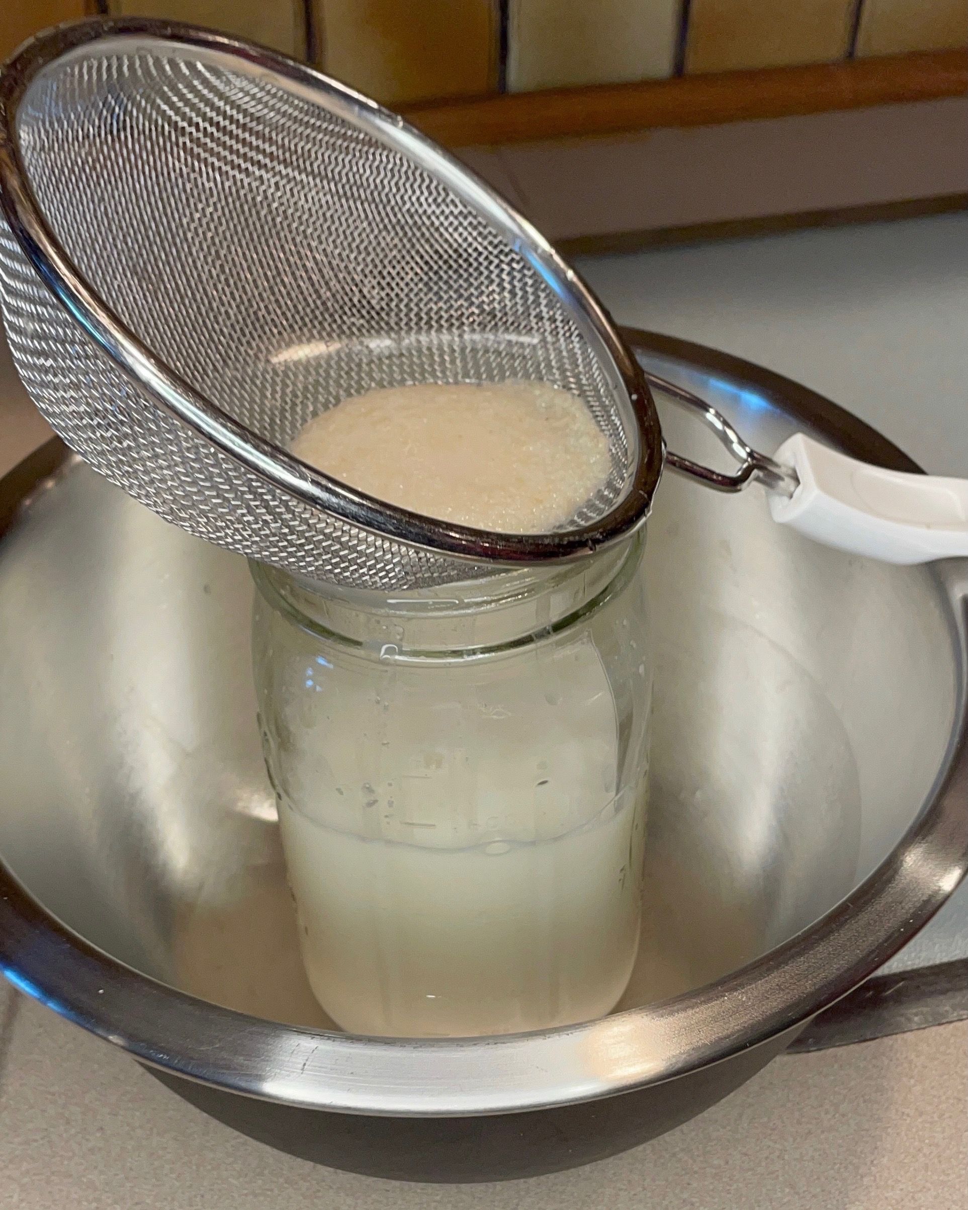 A mason jar of milk sitting inside of a steel bowl. A mesh strainer with caught rice pulp sits on the jar opening.