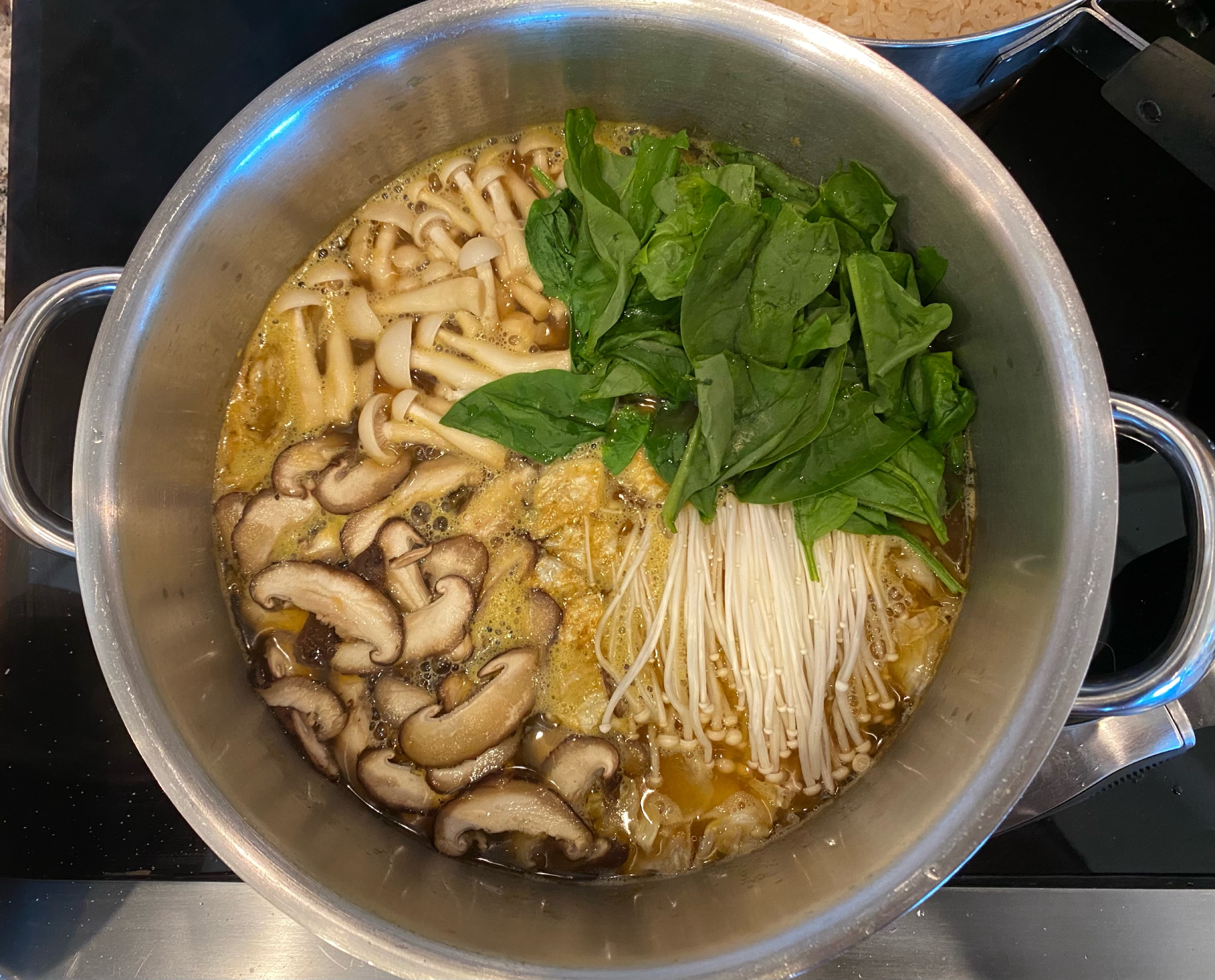 Overhead shot of a large soup pot. 3 types of mushrooms and a clump of spinach float on top in separate quartered sections. 