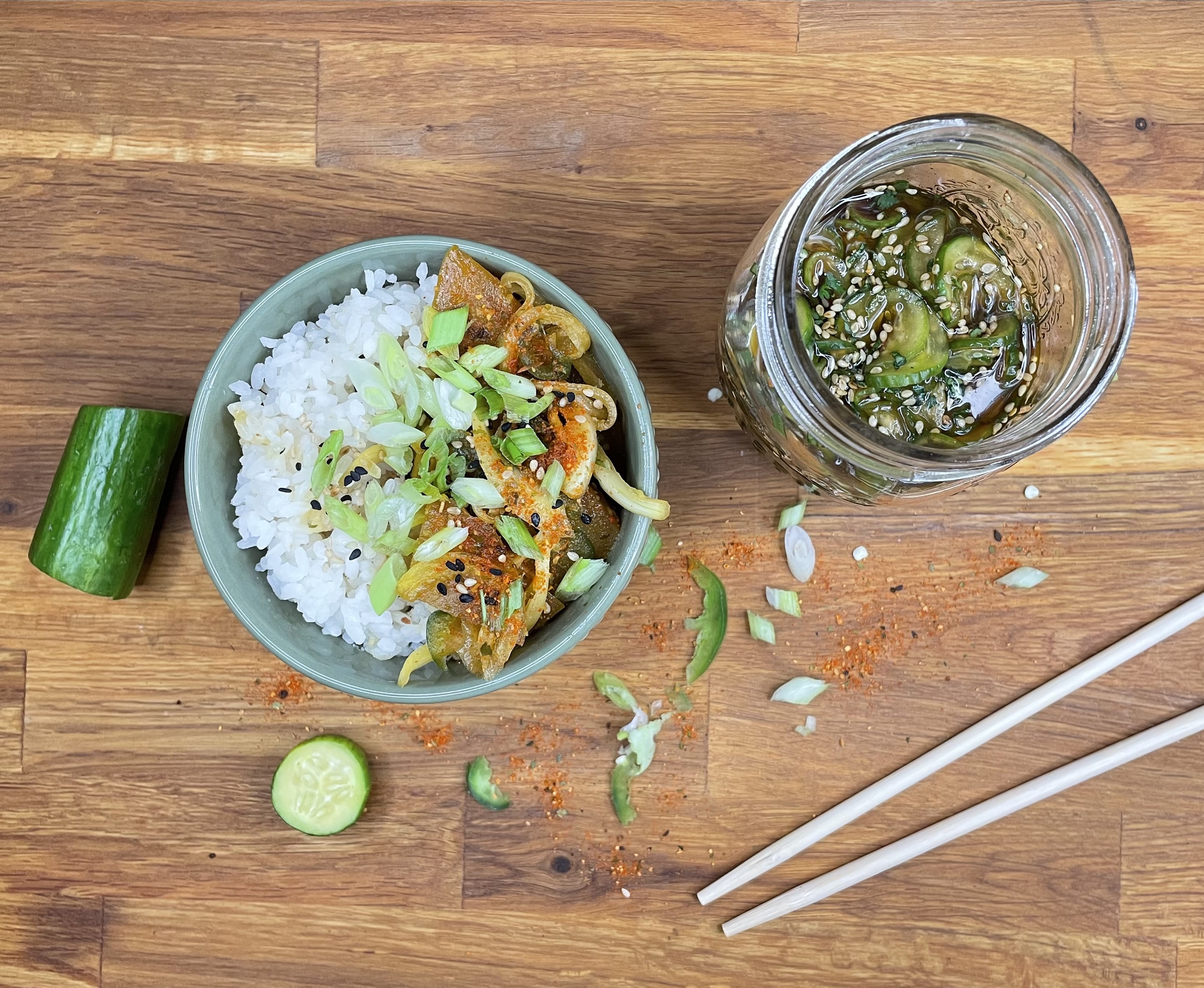 Overhead shot of a bowl of rice topped with golden beet poke, scallions, and sesame seeds. A jar with pickled cucumbers is beside the bowl. Chopsticks, jalapeños, scallions, lime, and cucumbers scattered are on the countertop 
